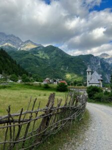 A scenic rural landscape in the Albanian Alps, featuring a traditional wooden fence, a small church, a village nestled among mountains, and a field of wildflowers under a cloudy sky.