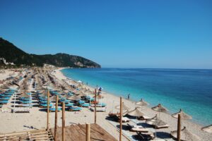 A beautiful sandy beach with clear blue water and mountains in the background. There are many sun umbrellas and lounge chairs set up on the beach.