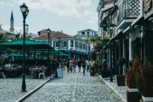 A cobblestone street in Korçë, Albania, lined with outdoor cafés, green umbrellas, and traditional buildings in the background.